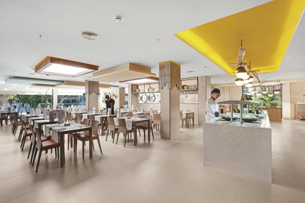 Mūsdienīgs restorāns ar elegantu interjeru, kurā redzami koka galdi un krēsli, un darbinieks, kas strādā pie bufetes. Gaismas elementi un zaļa auga klātbūtne rada patīkamu atmosfēru.