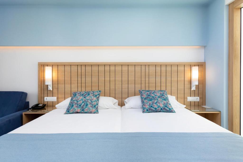 Mūsdienīgs guļamistabas interjers ar divām gultām, dekorētām ar spilveniem ar tropu rakstu, un gaiši zilu sienu. Blakus atrodas dīvāns un galda lampa.