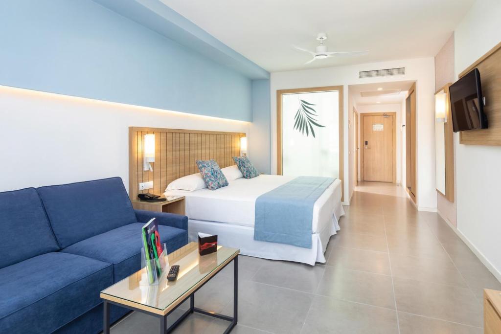 Mūsdienīga viesnīcas numura iekārtojums ar zilu dīvānu, gultu un koka elementiem, kas rada patīkamu atmosfēru. Numurā ir arī televizors un galdiņš ar piederumiem.