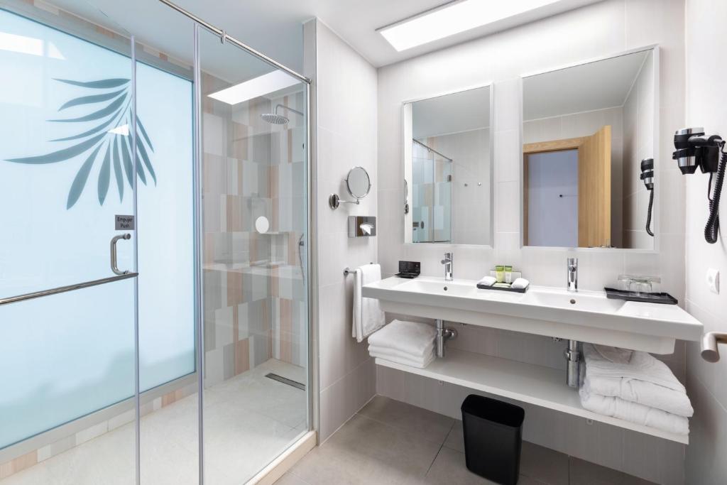 Mūsdienīgs vannas istabas interjers ar stikla dušu un divām spoguļu virsmām, kas papildināts ar zaļiem aksesuāriem un baltām dvieļiem.