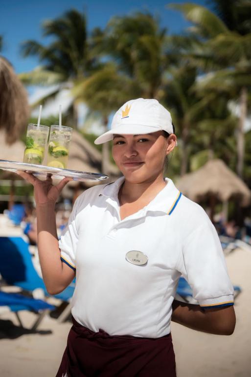 Pavedienu pasniedzēja pludmalē nes pasniegšanas paliktni ar kokteiļiem, kamēr apkārt ir palmām apstādīta vide.