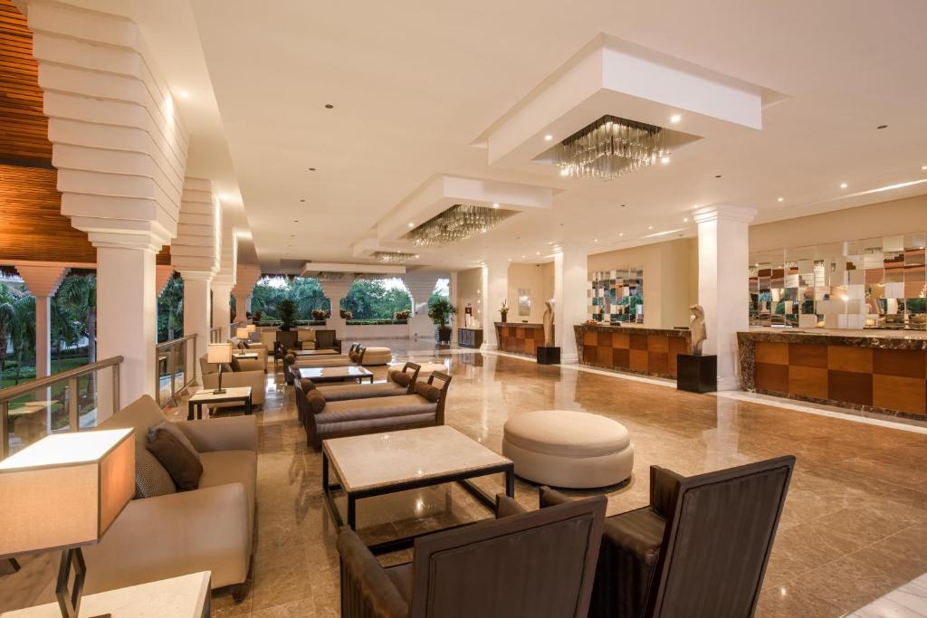 Mūsdienīgs viesnīcas vestibils ar elegantiem dīvāniem un galdiem, apgaismots ar stilīgām lampām un greznu griestu lampu. Fons veido zaļi augi un plašs skats uz āru.