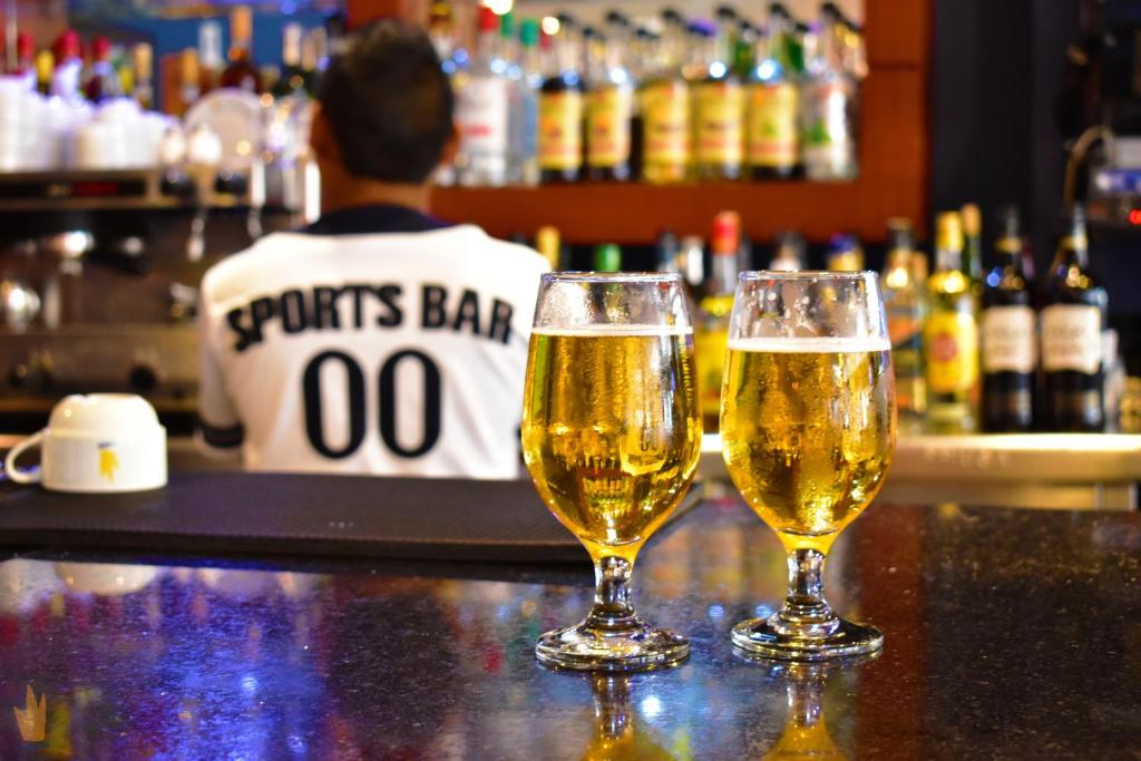 Divas glāzes ar dzidru alu atrodas uz bāra letes, fonā redzams bārmenis un alkohola dzērienu plaukti.