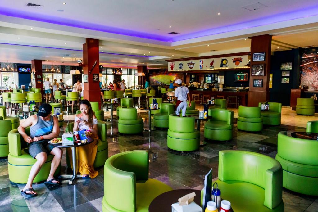 Mūsdienīga restorāna iekārtojums ar zaļām krēsliem, cilvēki bauda maltītes un dzērienus. Gaisotne ir dzīvīga, ar sporta tematikas dekorācijām un lielu bāru.