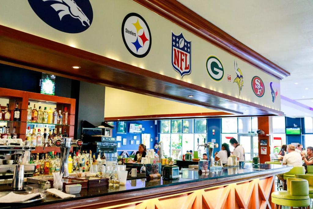 Bārs ar NFL komandu logotipiem uz sienas un plašu dzērienu izvēli. Apmeklētāji izbauda atmosfēru un sporta pārraides.