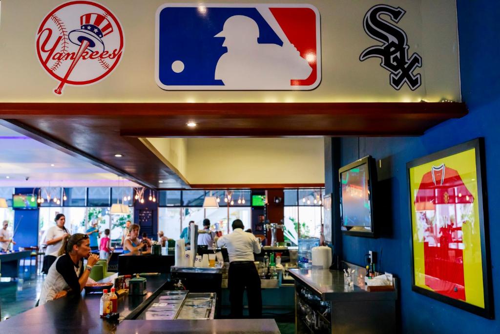 Restorāna interjers ar MLB logotipu un komandu emblēmām, tostarp Yankees un White Sox, kā arī apmeklētājiem, kas izbauda atmosfēru.