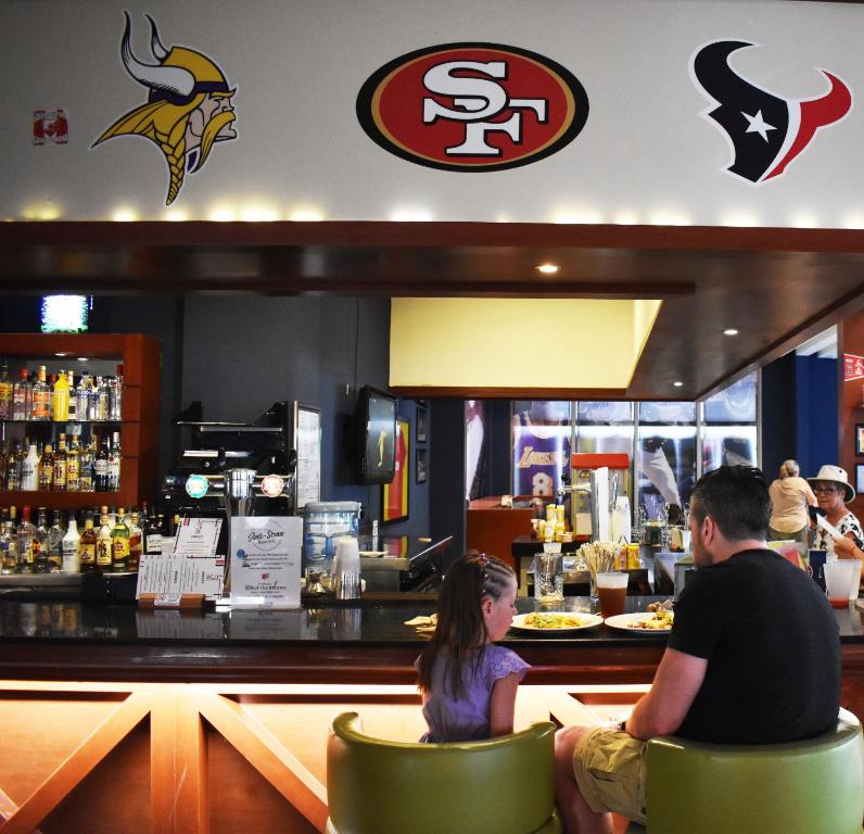 Bārs ar sporta tematikas dekorācijām, kur pie letes sēž pieaugušais un bērns, baudot ēdienus un dzērienus. Virs bāra redzamas NFL komandu emblēmas.