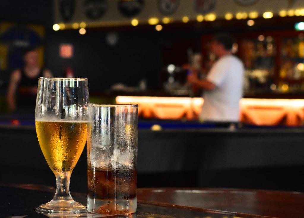 Divi dzērieni bāra galda priekšplānā, vienā pusē ir dzidrs alus, otrā pusē - kokteiļa glāze ar ledu. Fons ir izplūdis, bet redzams bārs un cilvēks, kas apkalpo.