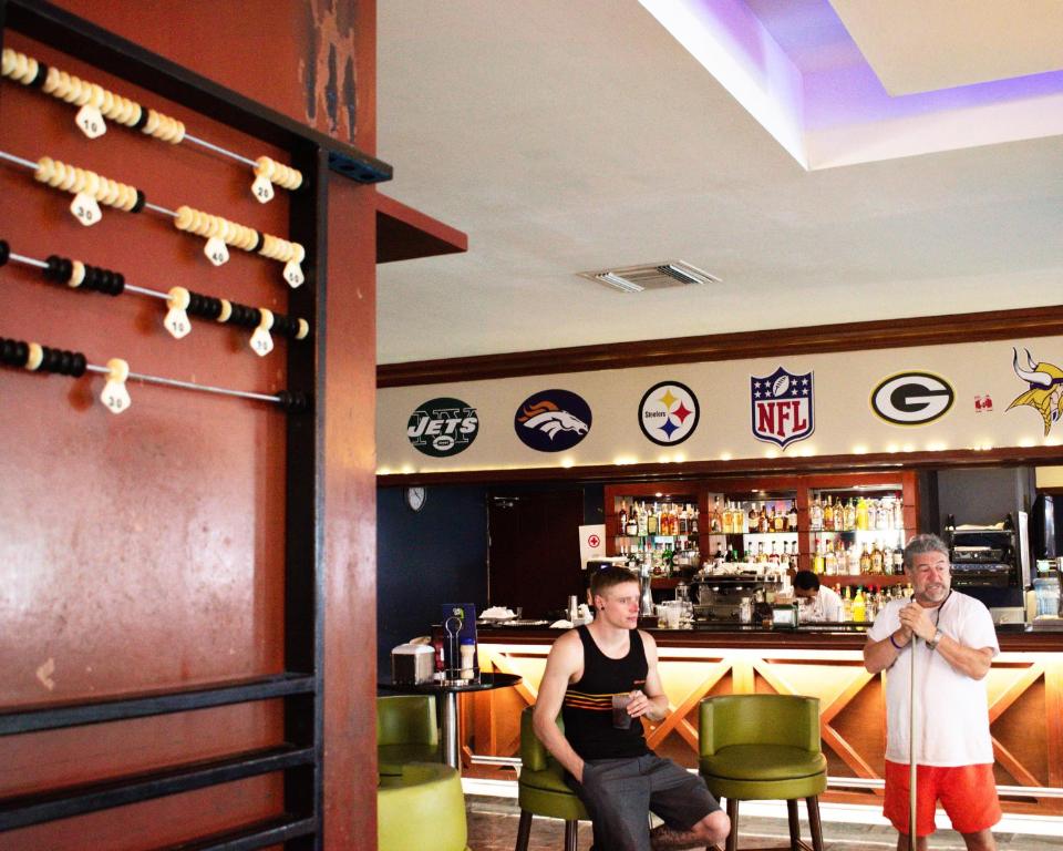 Bārs ar sporta logoem un cilvēkiem, kas atpūšas pie galda. Interjerā redzama skaitļu skaitītāja un bāra zona ar dzērieniem.