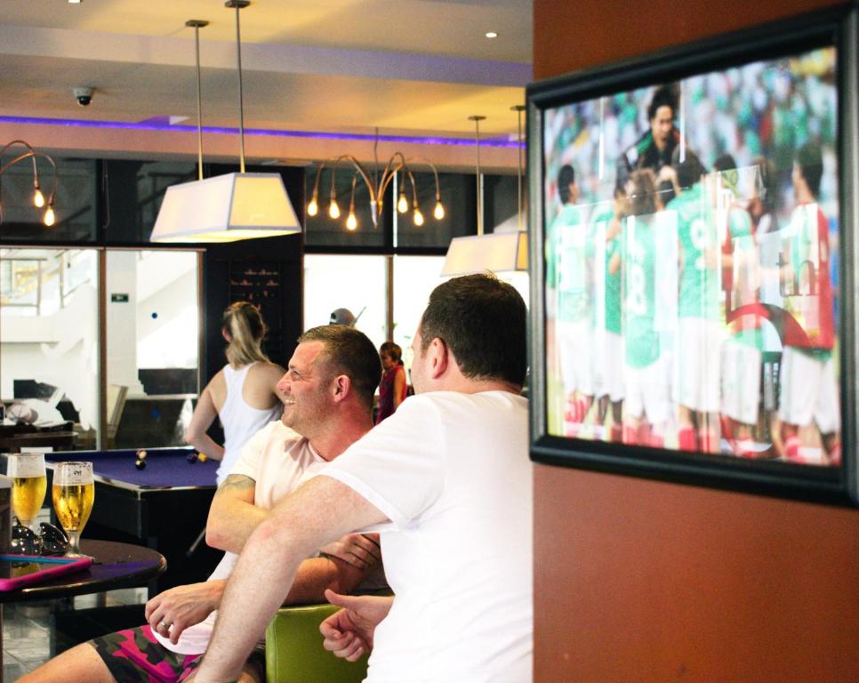 Divi vīrieši relaksējas bārā, baudot dzērienus un skatoties uz futbolu, kas redzams attēlā. Fons ietver bāra interjeru un citas viesus.