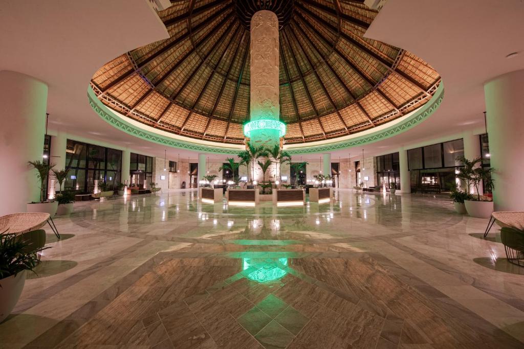 Mūsdienīgs viesnīcas vestibils ar iespaidīgu apaļu jumtu, centrā esošu kolonnas struktūru un zaļganu apgaismojumu, radot elegantu un relaksējošu atmosfēru.