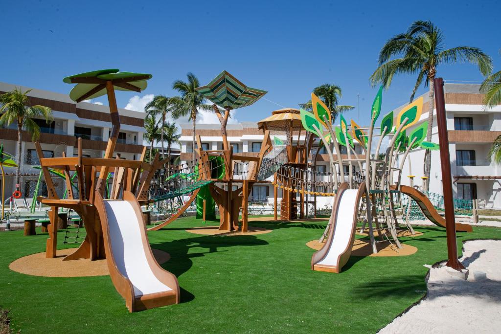Bērnu laukums ar koka konstrukcijām, slīdkalniņiem un virvju tiltu, ielikts zaļajā zālienā, ar palmām fonā un modernu viesnīcu.