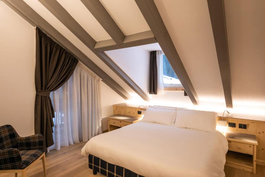 Mūsdienīga guļamistaba ar slīpiem griestiem, baltu gultu un koka mēbelēm, kurā ir ērts krēsls un aizkari. Telpa ir gaiša un mājīga, radot relaksējošu atmosfēru.