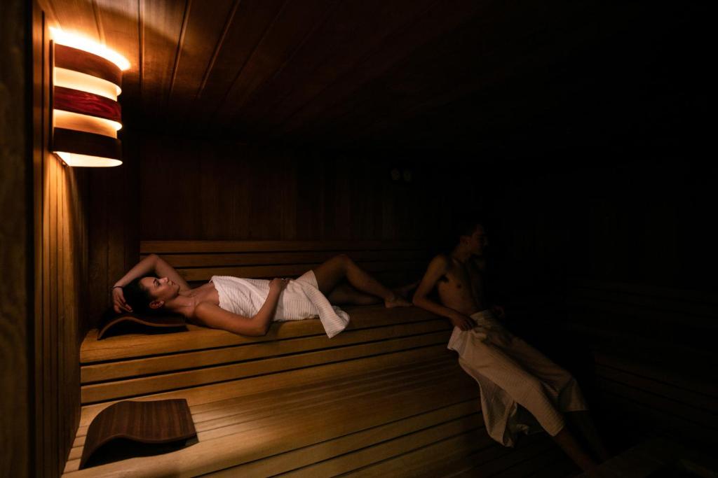 Divi cilvēki relaksējas koka pirtī, viens guļ uz soli un otrs sēž. Apkārt ir siltas gaismas un koka apdare, radot mierīgu atmosfēru.