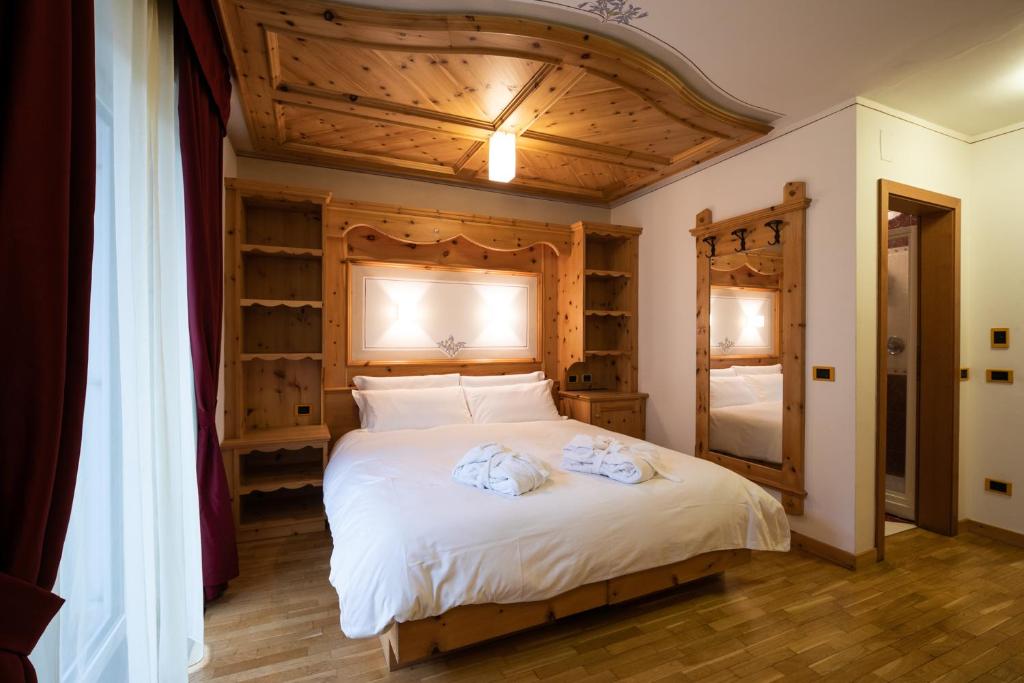 Mūsdienīgs guļamistabas interjers ar koka mēbelēm un siltu apgaismojumu, piedāvājot mierīgu un mājīgu atmosfēru. Gultā ir balta veļa, un telpā ir liels spogulis ar dekoratīvām detaļām.