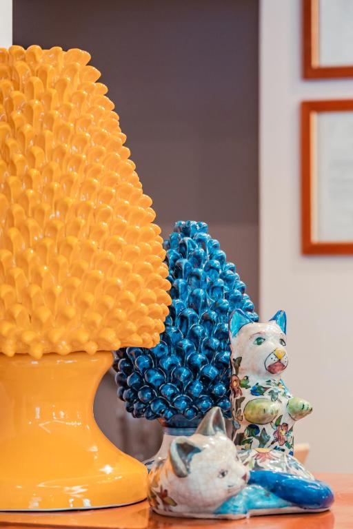 Krāsainu keramikas dekori, tostarp dzeltena lampa ar izteiksmīgu dizainu, zila figūra un divas porcelāna kaķu figūras, kas piešķir dzīvīgu atmosfēru.