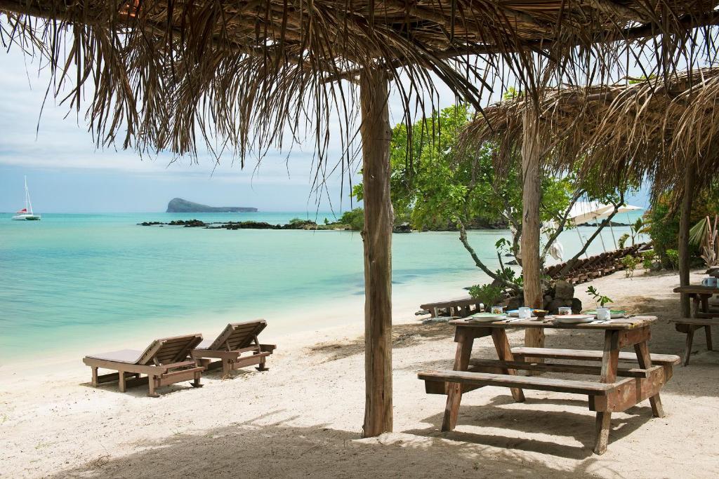 Pludmales ainava ar divām atpūtas krēslām, koka galdu un krēsliem, kas izvietoti zem jumta, ar skatu uz mierīgu jūru un nelielu salu tālumā.