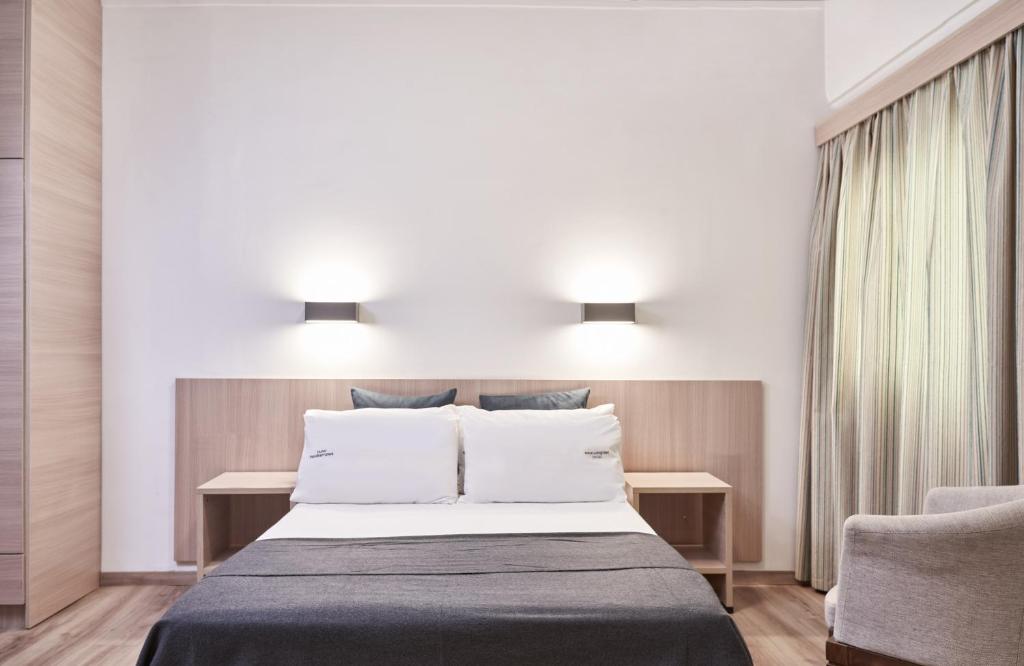 Mūsdienīgs guļamistabas interjers ar ērtu gultu, koka mēbelēm un dabīgu apgaismojumu. Gultas veļā ir baltas un pelēkas krāsas akcenti, radot mierīgu atmosfēru.