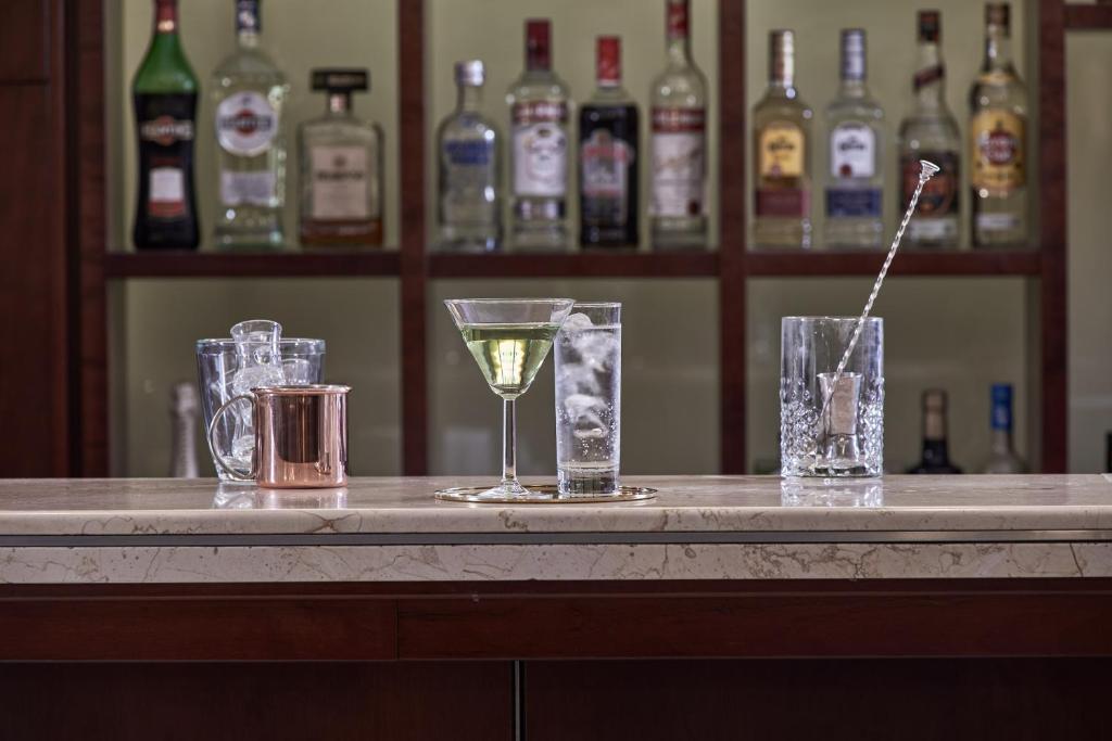 Eleganta bāra ainava ar dažādiem kokteiļu traukiem un dzērieniem, kas izvietoti uz marmora virsmas. Fons rotā ar dažādām alkohola pudelēm, radot viesmīlīgu atmosfēru.