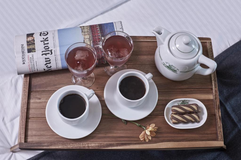 Kafijas un tējas komplekts uz koka paplātes, kopā ar divām dzērienu glāzēm un žurnālu. Blakus atrodas cepumi un zieds, radot relaksējošu atmosfēru.