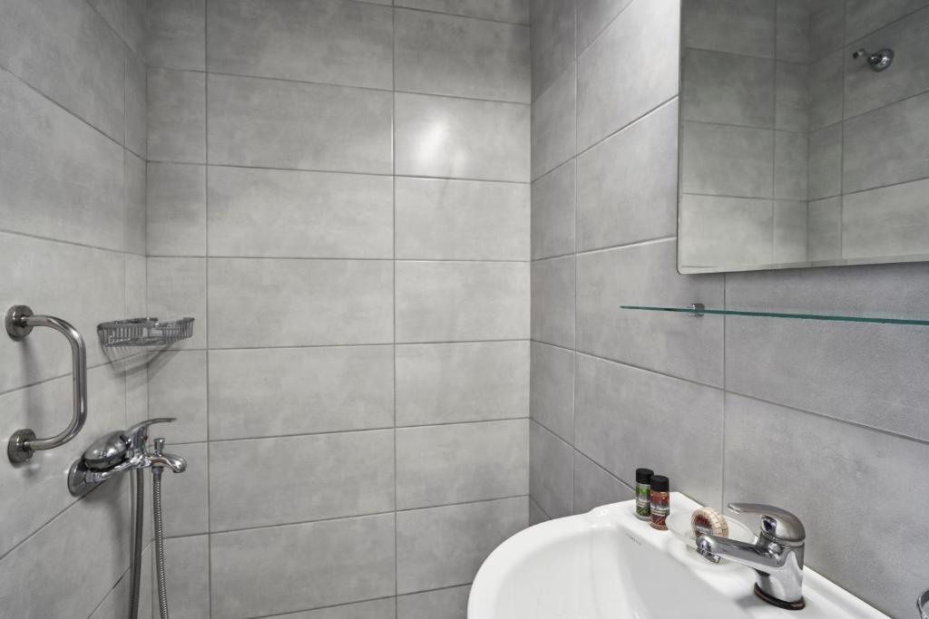 Mūsdienīgs vannas istabas interjers ar pelēkām flīzēm, dušas stūri un spoguli. Redzami higiēnas piederumi un hromētas santehnikas elementi.
