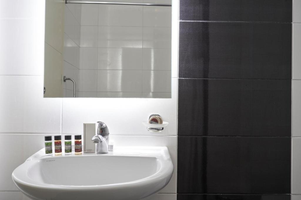 Mūsdienīga vannas istaba ar balto izlietni, spoguli un higiēnas produktiem, kas sakārtoti uz letes. Melna flīžu siena piešķir elegantu izskatu telpai.