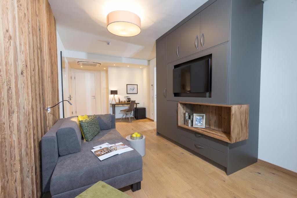 Mūsdienīgs viesistabas interjers ar pelēku dīvānu, koka akcentiem un televizoru. Telpā ir gaišs apgaismojums un stilīga dekorācija.