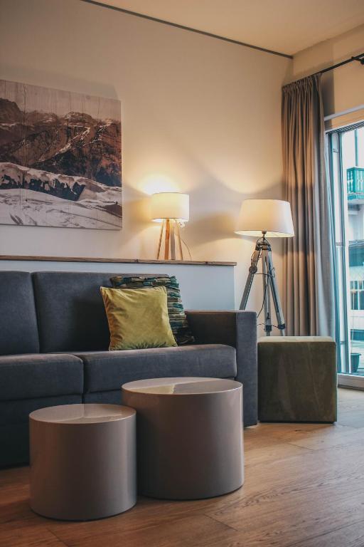 Mūsdienīgs viesistabas interjers ar pelēku dīvānu, dekoratīvu spilvenu, stilizētu gleznu uz sienas un dažādiem galdiem. Gaismas avoti rada siltu atmosfēru.