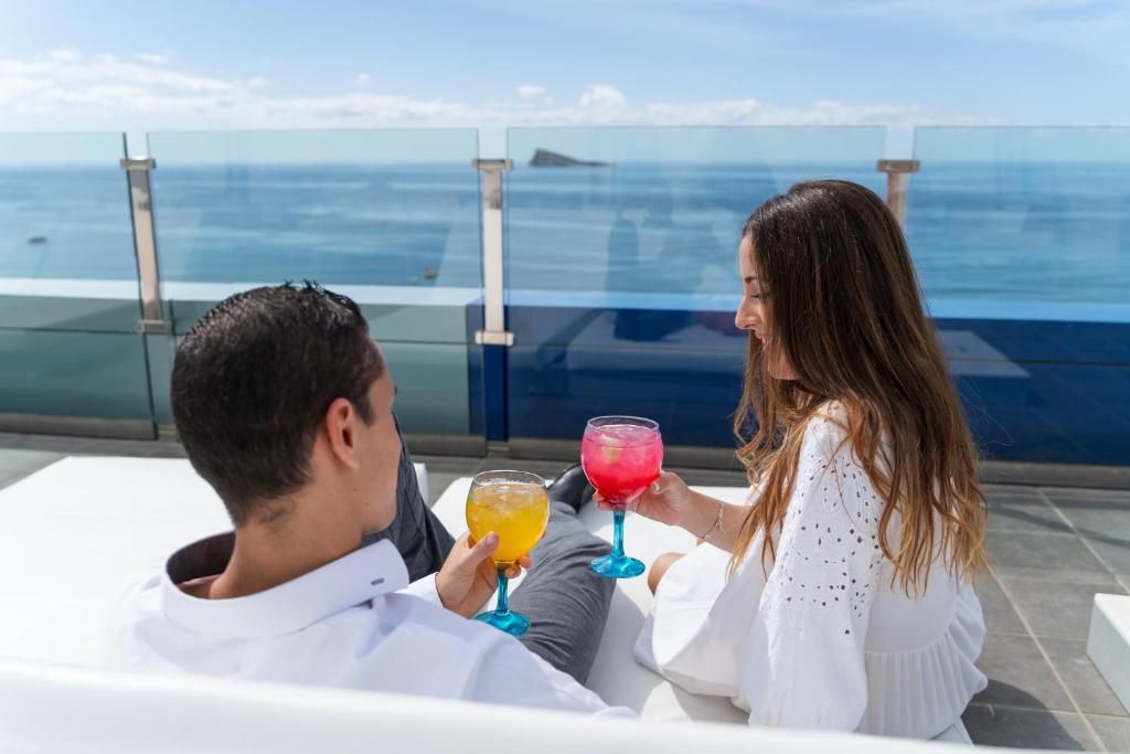 Pāris izbauda dzērienus uz balkona ar skatu uz jūru, sēžot komfortablās pozīcijās. Diena ir saulaina un skaidra, radot romantisku atmosfēru.