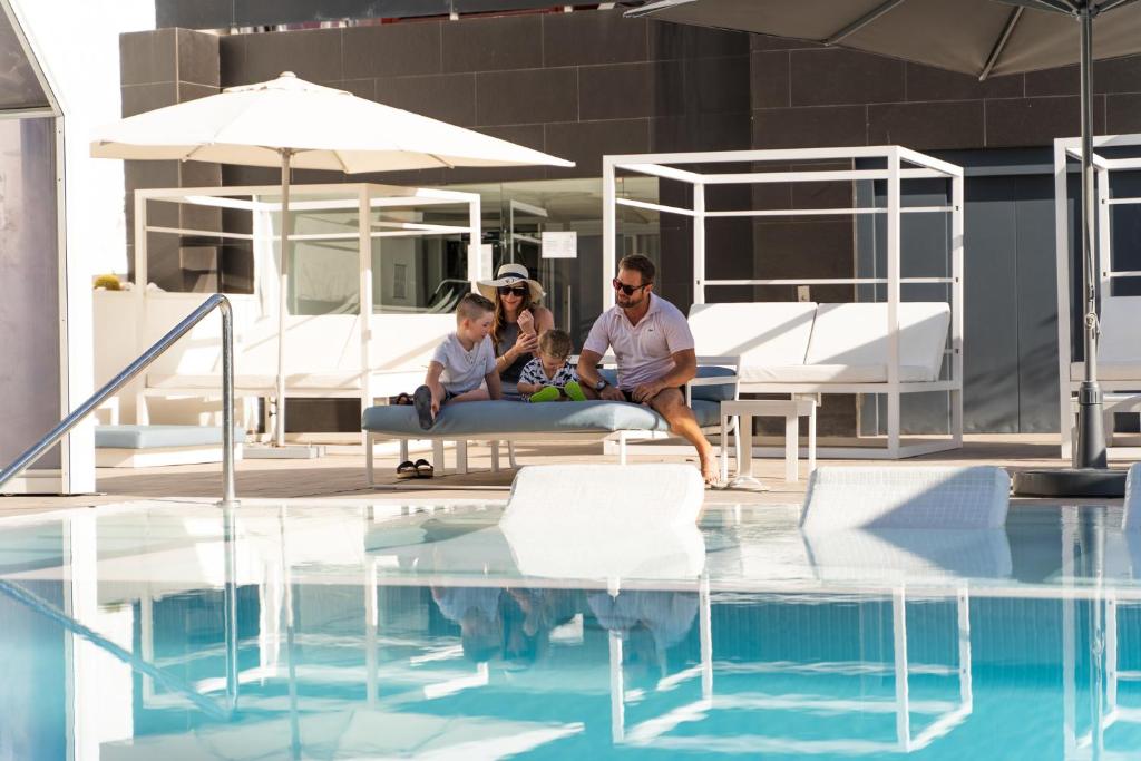 Ģimenes brīvdiena pie baseina, kur bērni un pieaugušie kopā atpūšas un izbauda sauli. Skats uz baseinu ar spoguļattēlu un modernām atpūtas zonām.