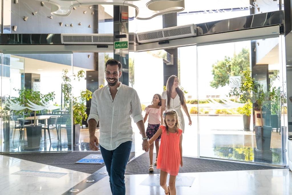 Ģimene ienāk modernā viesnīcā, priecīgi sveicot cits citu. Gaismas piepildītā telpā redzami augi un moderna arhitektūra.
