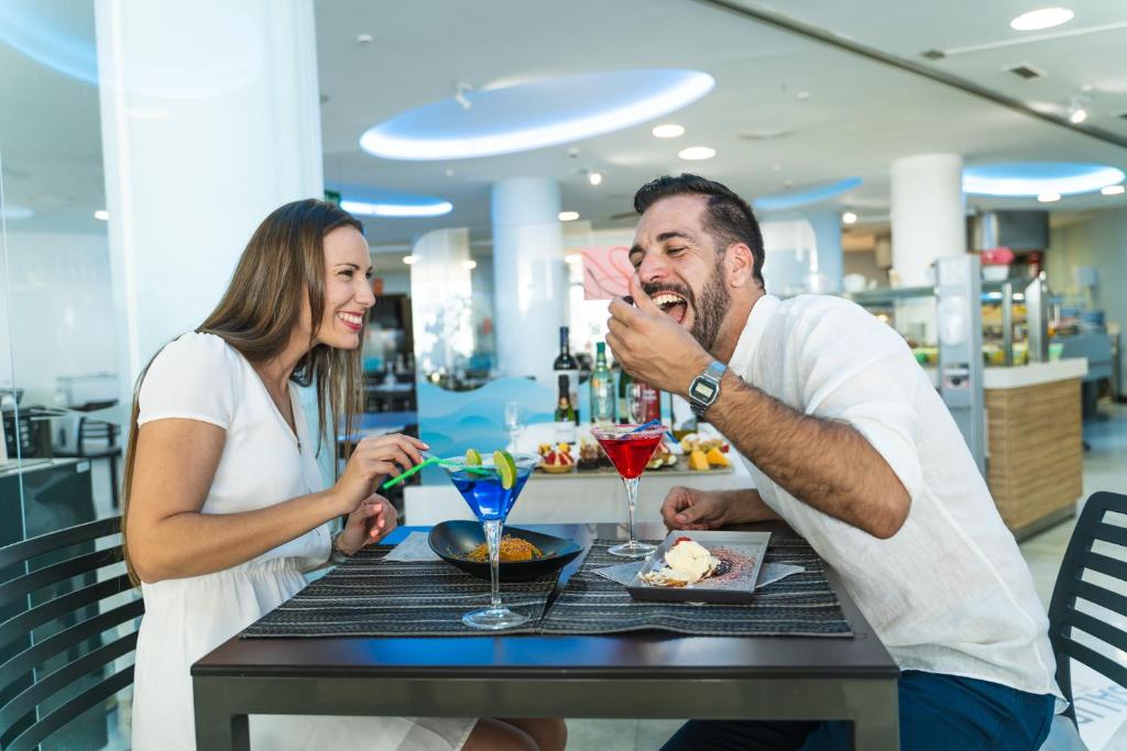 Divi cilvēki, vīrietis un sieviete, bauda maltīti restorānā, smejoties un sarunājoties, kamēr priekšā uz galda ir kokteiļi un ēdieni.