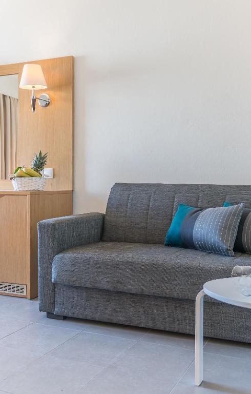 Mūsdienīgs viesnīcas numurs ar pelēku dīvānu, dekoratīvajām spilvenēm un koka kumodi, uz kuras atrodas augļu grozs un spogulis. Interjers ir gaišs un elegants.