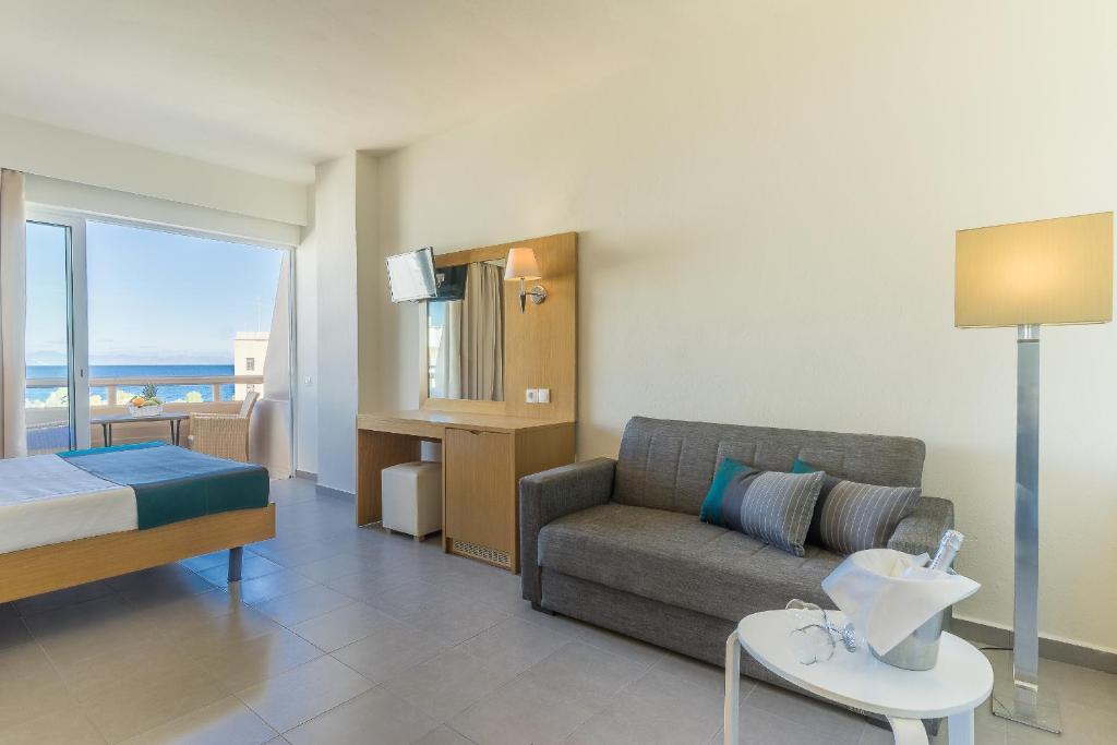 Mūsdienīga viesnīcas numura interjers ar skatu uz jūru, iekārtots ar ērtu dīvānu, televizoru un gaismas avotu. Numurs ir gaišs un plašs, radot relaksējošu atmosfēru.