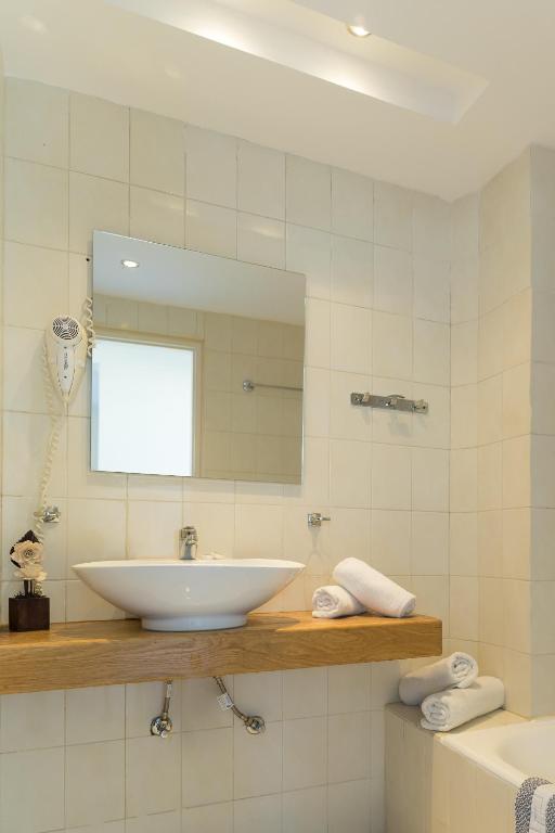 Mūsdienīgs vannas istabas interjers ar gaišām flīzēm, koka plauktu un elegantu izlietni. Uz plaukta redzami dvieļi un dekoratīvs elements.