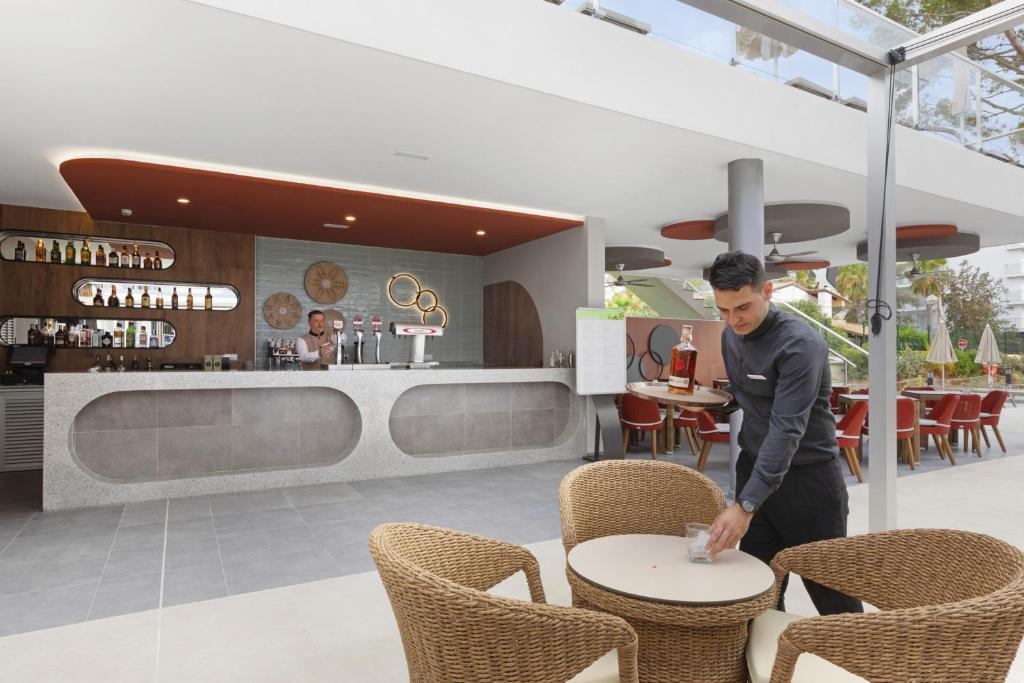 Mūsdienīgs bārs ar jauku interjeru, kur viesmīlis apkalpo klientus, kamēr otrais darbinieks strādā aiz bāra. Redzami eleganti krēsli un galdi, kas veido patīkamu atmosfēru.