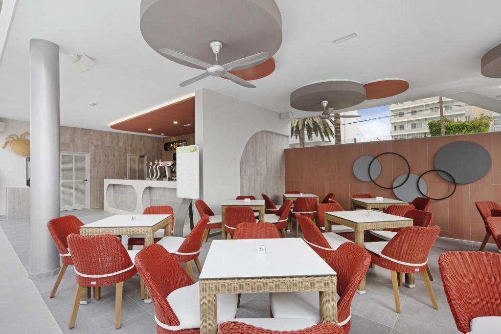 Moderns restorāns ar sarkaniem krēsliem un baltiem galdiem, kurā ir skaidrs interjers un lieli logi, kas nodrošina dabisko apgaismojumu. Sienā redzami dekoratīvi elementi un bārs.