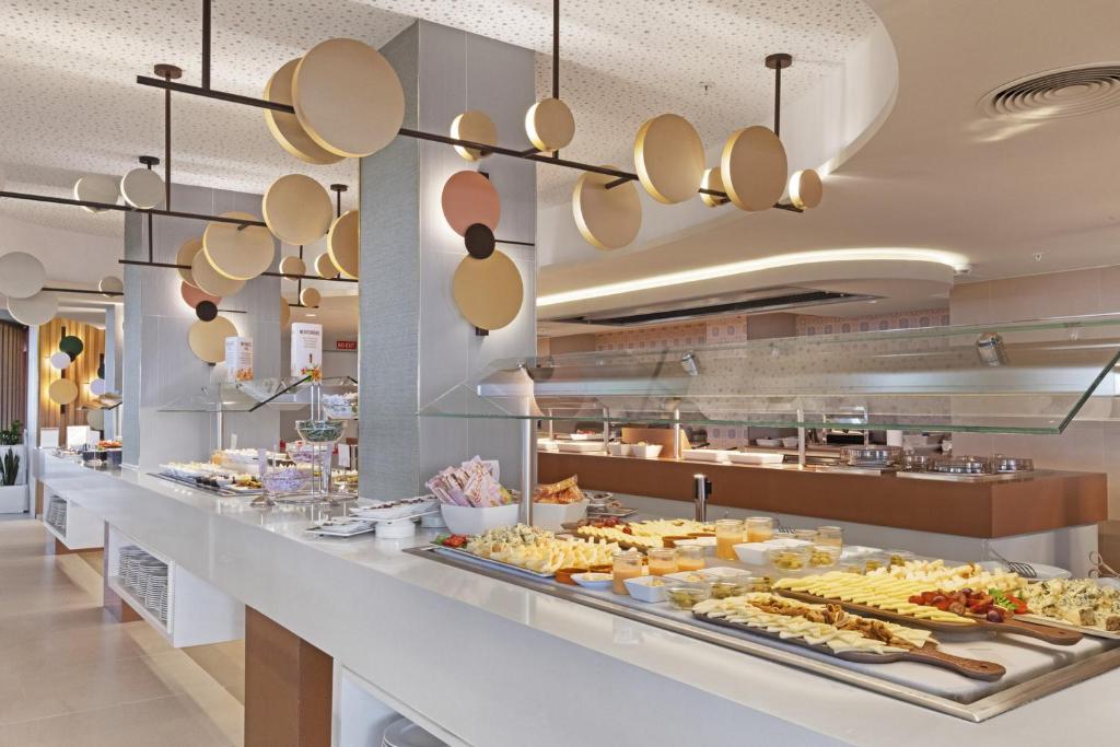Moderns restorāns ar plašu bufeti, kurā redzams dažādu ēdienu klāsts un stilīgas gaismas ar apaļām formām. Eleganta interjera dizains rada patīkamu atmosfēru.
