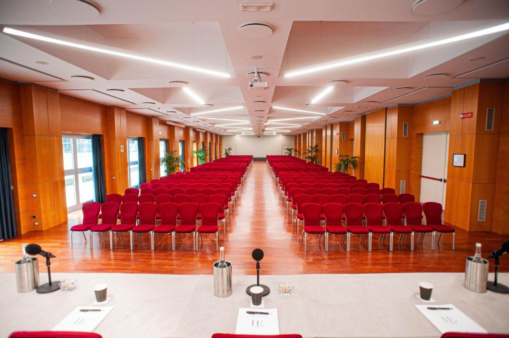 Konferences telpa ar sarkaniem krēsliem, kas sakārtoti rindās gar garu koka grīdu. Telpā ir lieli logi un augu dekorācijas, radot profesionālu un viesmīlīgu atmosfēru.