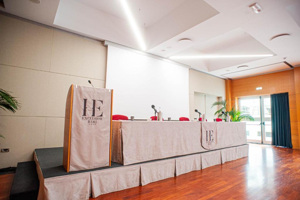 Konferenču zāle ar podiumu, uz kura ir mikrofoni un ūdens, gaida prezentāciju. Fons ir gaišs, ar lielu ekrānu un logiem, caur kuriem redzama zaļa auga daļa.