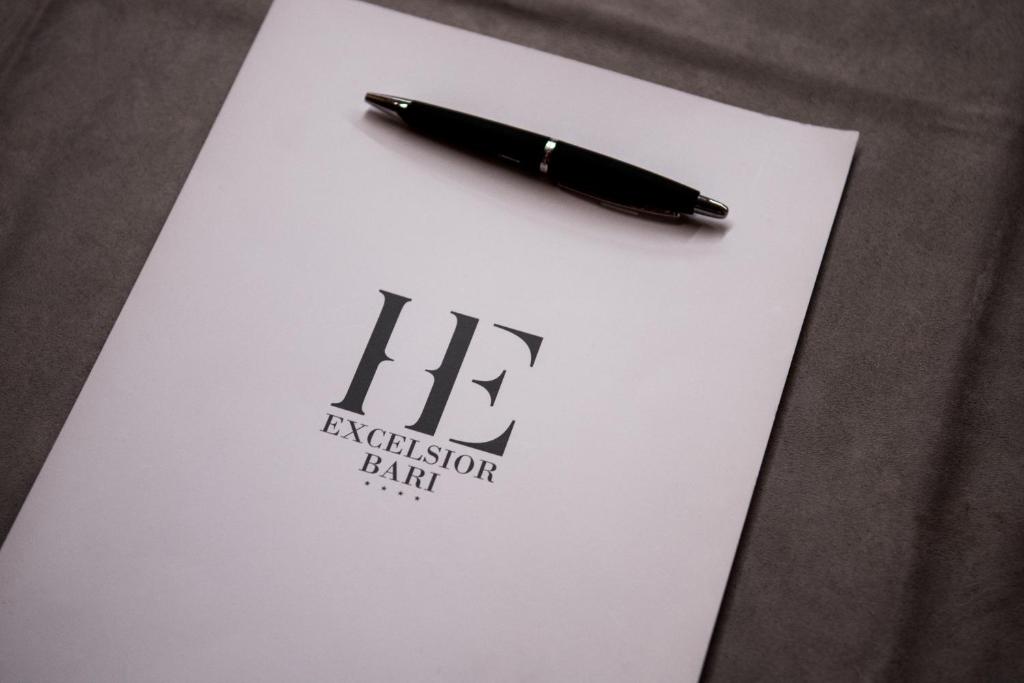 Baltā papīra lapa ar melnu logotipu "Excelsior Bari" un melnu pildspalvu uz pelēka fona.