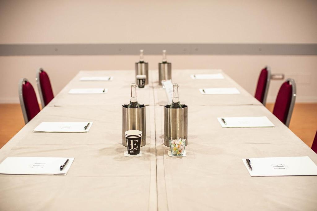 Konferenču telpa ar garu galdu, kas sagatavots sanāksmēm, uz galda ir ūdens pudeles, kafijas tases un piezīmju lapas. Sarkanās krēslus ap galdu veido profesionālu atmosfēru.