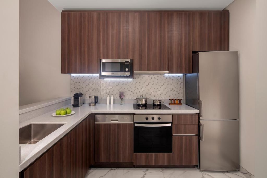 Mūsdienīga virtuve ar koka skapjiem, sudraba ledusskapi un siltu apgaismojumu, kurā redzami virtuves piederumi un zaļie āboli uz galda.