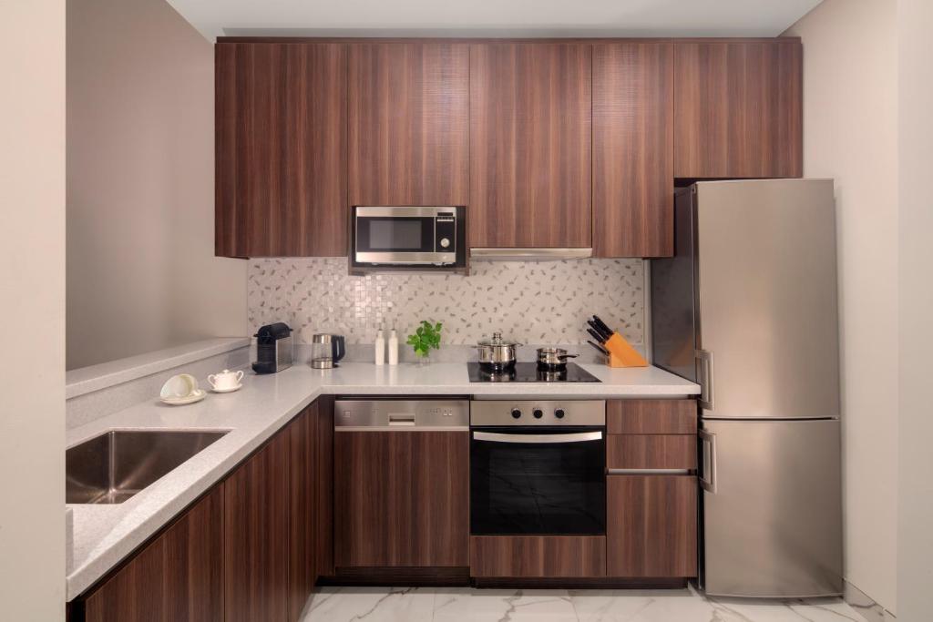 Mūsdienīga virtuve ar tumšu koka skapīšu dizainu, sudraba ledusskapi un modernu virtuves tehniku. Virsma ir no gaiša akmens, un ir redzamas arī dažas virtuves piederumu detaļas.