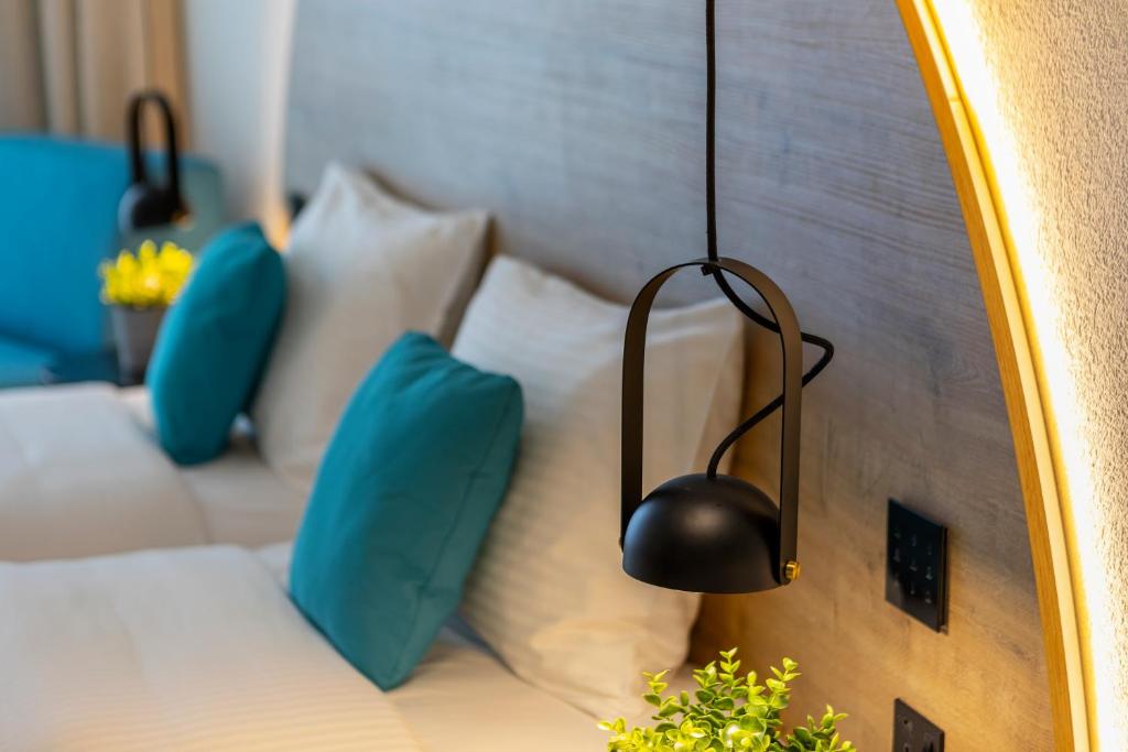 Mūsdienīgs guļamistabas interjers ar divām gultām, melnu piekaramo lampu un spilgtiem ziliem spilveniem. Aiz guļamistabas gultām redzama arī dekoratīva auga kompozīcija.