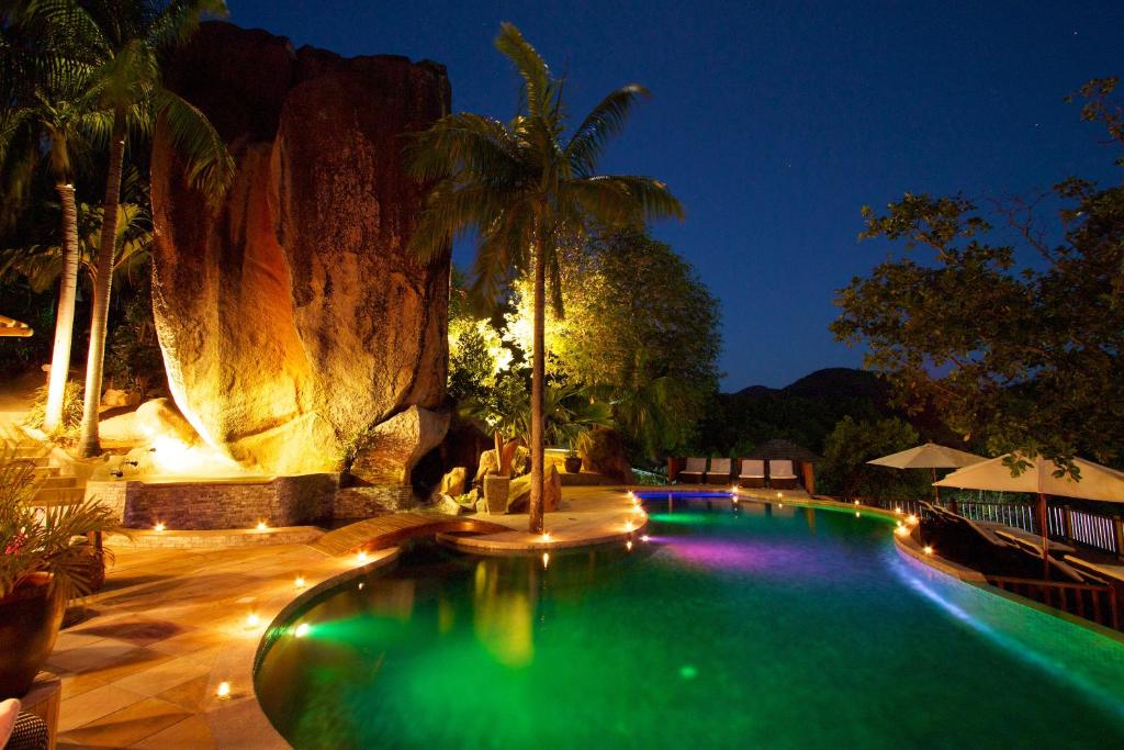 Eksotiska nakts ainava ar baseinu, kas apgaismots ar krāsainām gaismām, un apkārt esošajām palmu koku un akmeņu struktūrām, radot mierīgu atmosfēru.