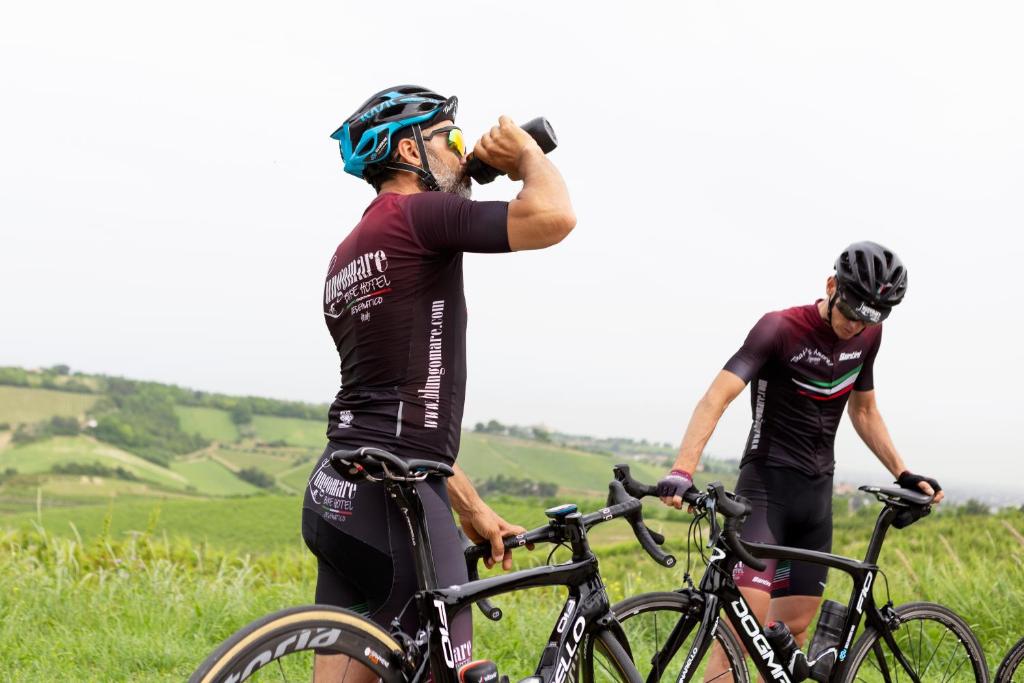 Divi riteņbraucēji atpūšas kalnu ainavā, viens no viņiem dzer ūdeni, bet otrs pārbauda velosipēdu. Fons ir zaļa daba ar viļņainiem laukiem.