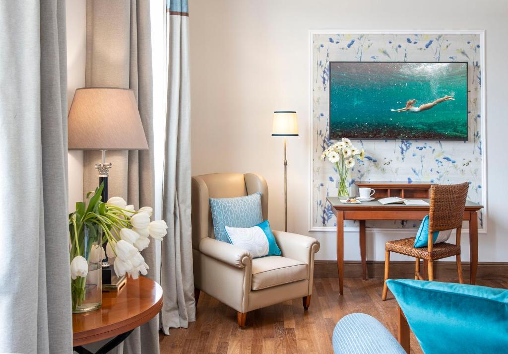 Mūsdienīgs viesnīcas numurs ar elegantu apdari, kas ietver gaismas toņu aizkarus, mājīgu atpūtas krēslu, ziedu kompozīciju un gleznainu ainavu uz sienas. Ideāla vieta relaksācijai un iedvesmai.