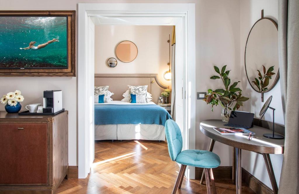 Mūsdienīgs viesnīcas numurs ar elegantu interjeru, kas ietver zilo gultas pārklāju un mākslas darbu, kurā attēlota peldētāja. Telpā ir arī rakstāmgalds un ziedi, radot patīkamu atmosfēru.