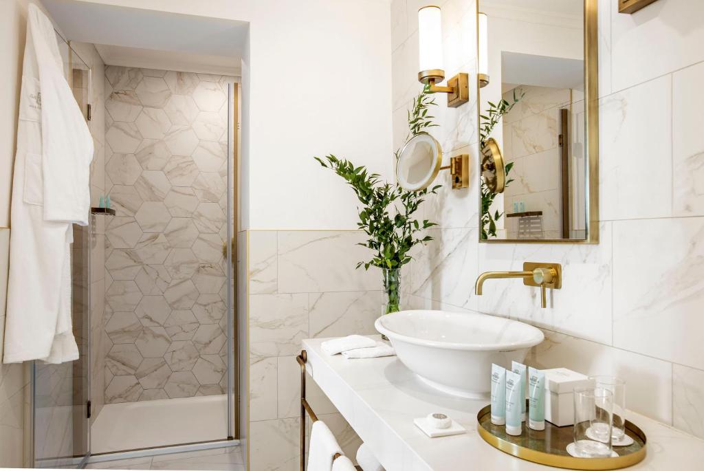 Mūsdienīgs vannas istabas interjers ar marmora flīzēm, elegantu izlietni un augu dekoru. Aiz stikla durvīm redzama duša, savukārt uz galda novietoti higiēnas līdzekļi.